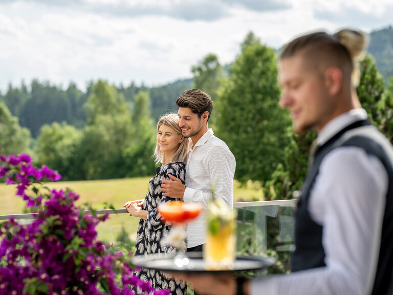 Ein verliebtes Paar beim gemeinsamen Getränk auf der Sonnenterrasse des Wellnesshotels Mooshof im Bayerischen Wald mit Blick auf die Naturkulisse.