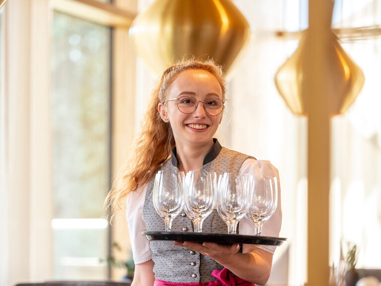 Eine Service Mitarbeiterin für das Restaurant im Hotel Mooshof in Bayern lächelt glücklich mit Tablet in der Hand.